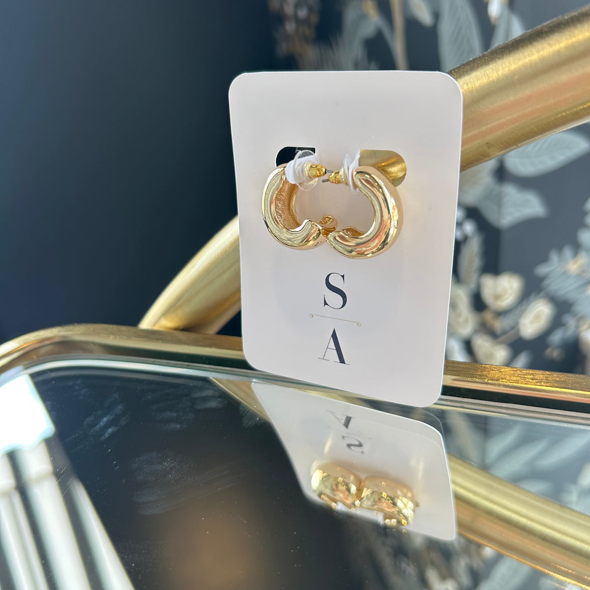Gold hoop earrings on a white card with 'S&A' branding, displayed against a reflective surface.