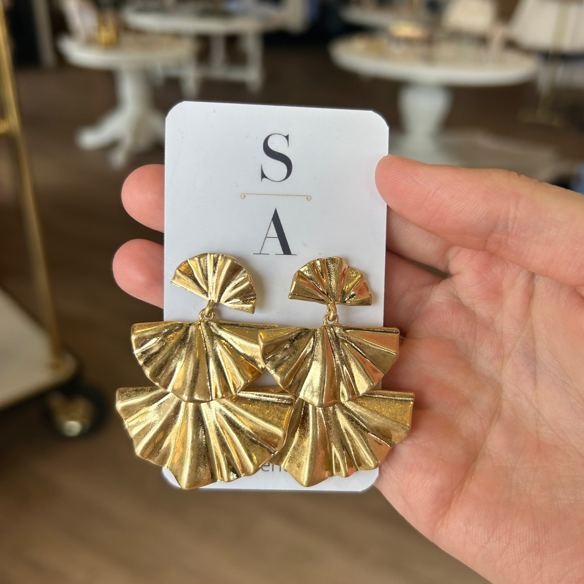 Gold fan-shaped earrings on a white card held by a hand with a blurred indoor background.