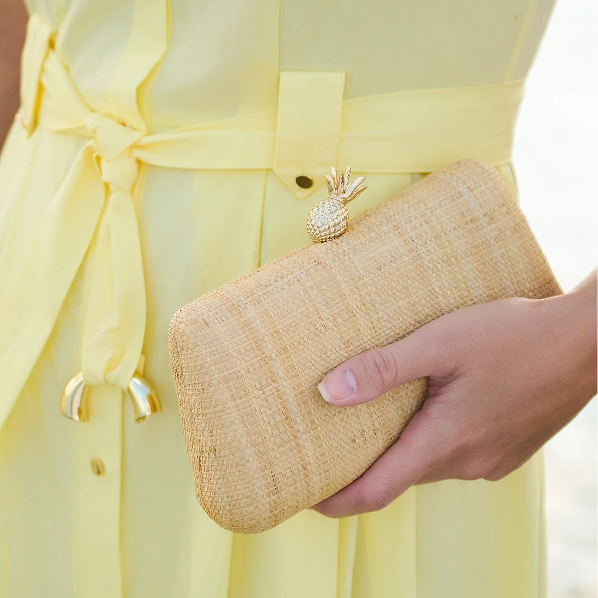 Person wearing a yellow dress holding a woven clutch with a pineapple charm.