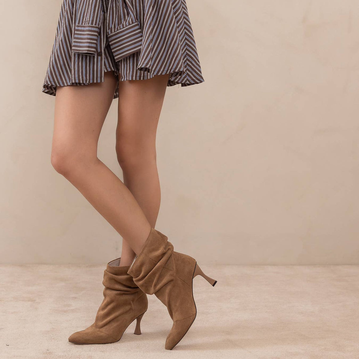 A model's legs wearing a gray and black striped ruffled mini skirt with a tie waist, paired with brown suede ankle boots, against a neutral beige background.