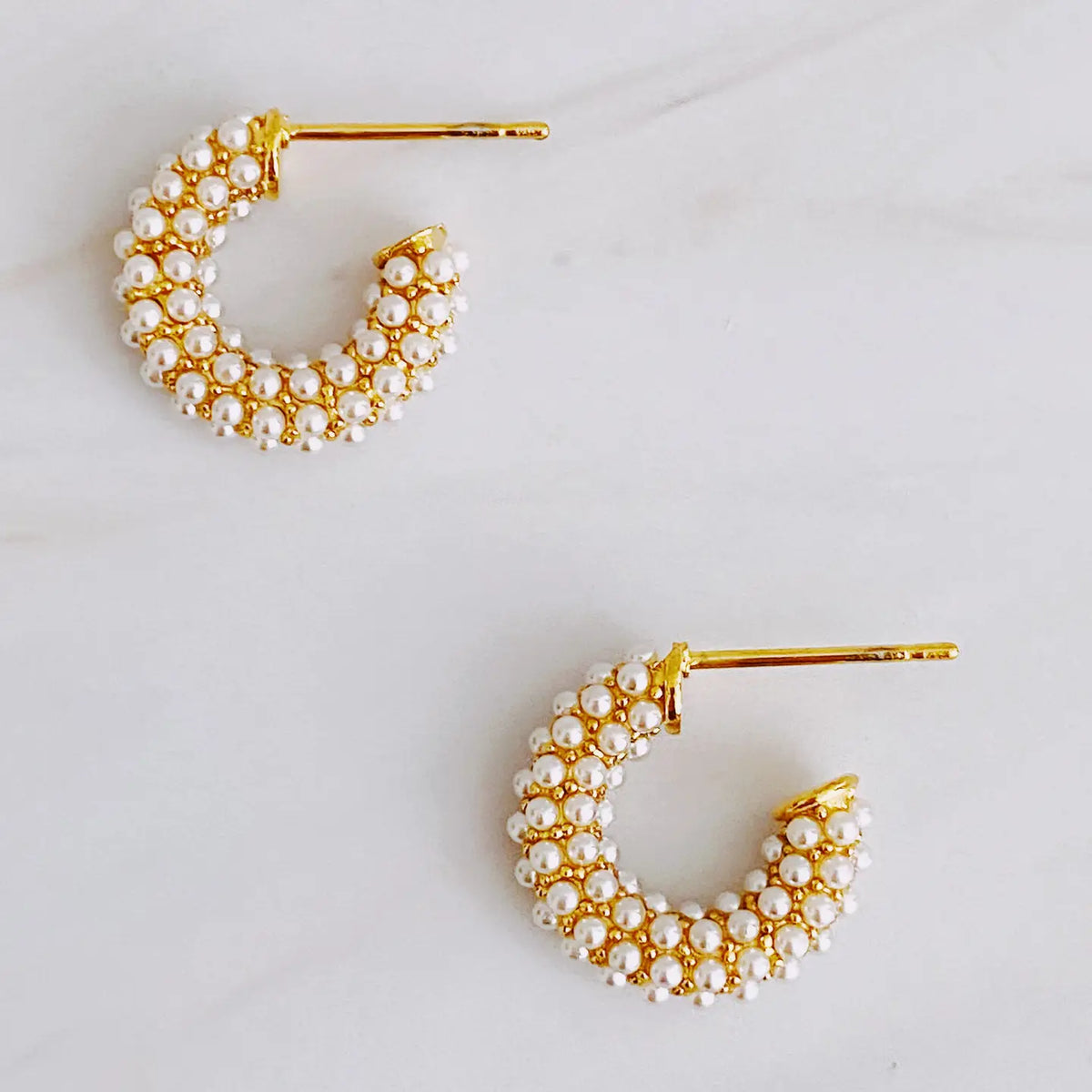 A pair of gold crescent-shaped hoop earrings adorned with small white pearls, displayed on a light marble surface.