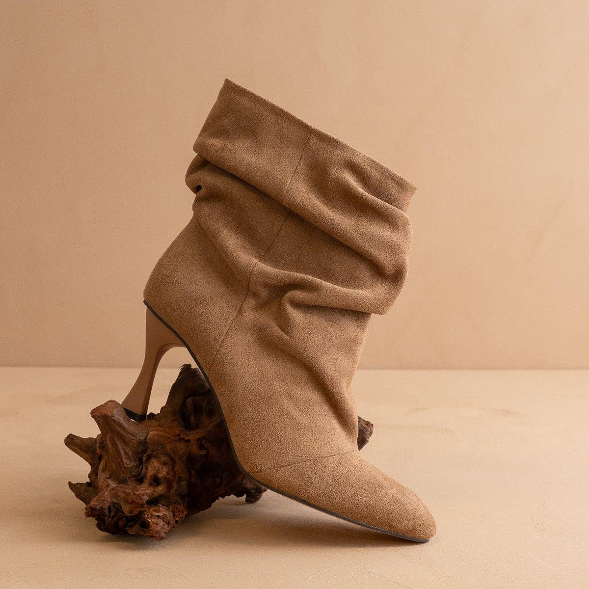 A single taupe-brown suede ankle boot with a pointed toe and a sculptural high heel, styled on a piece of dark wood against a warm beige background.