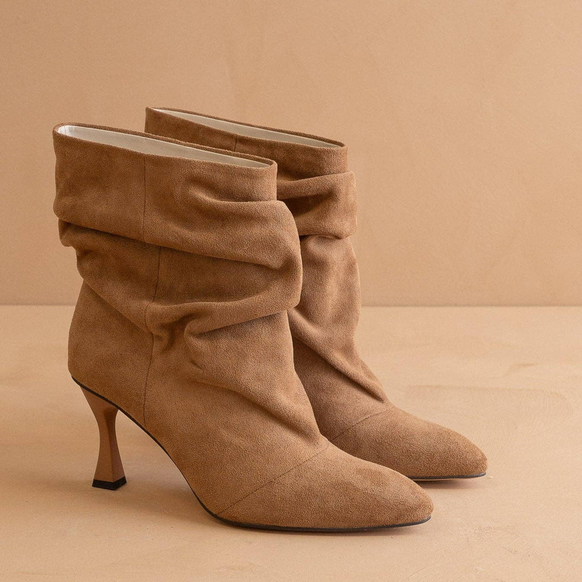 A pair of taupe-brown suede ankle boots with a slouchy draped design, pointed toes, and slim kitten heels, set against a warm beige background.