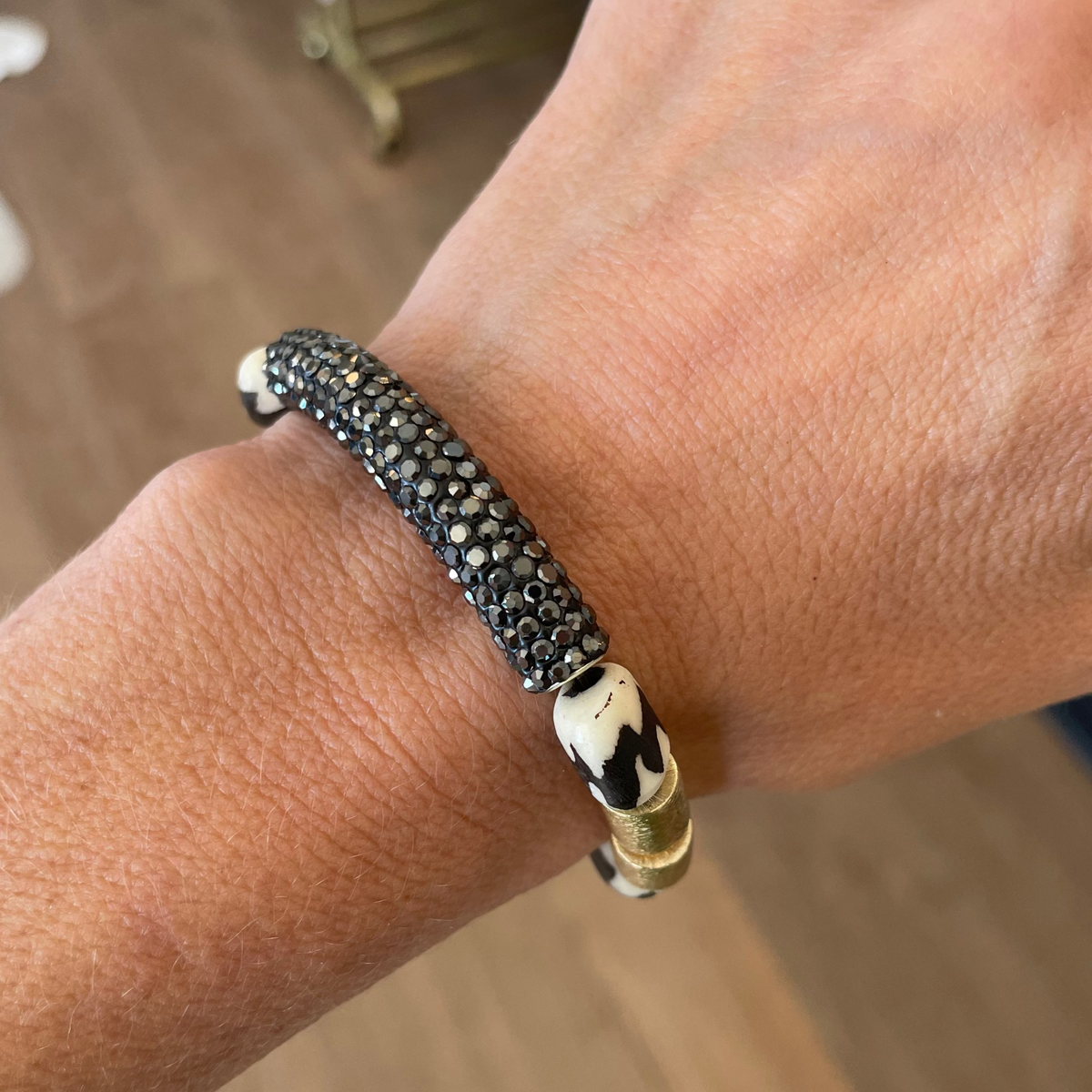 Close-up of a wrist wearing a black and gold bracelet with a blurred indoor background.