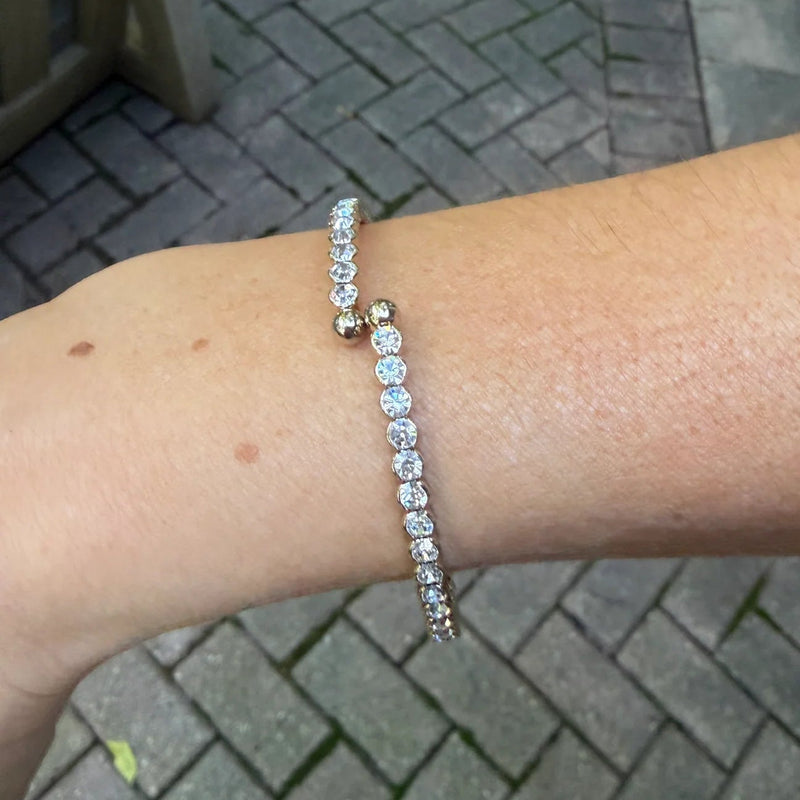 A close-up of a person's wrist wearing a delicate bracelet with a line of clear round-cut stones and a small gold charm, photographed outdoors on a paved surface.