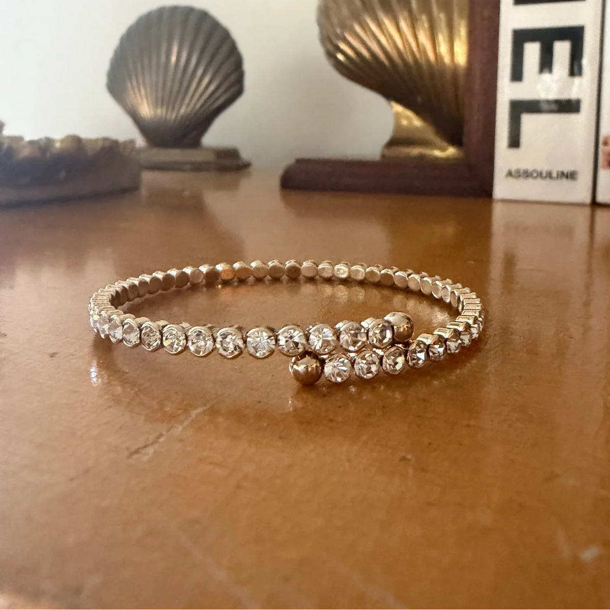 A delicate gold bangle bracelet with a row of clear round-cut stones and a small gold bead accent, resting on a wooden tabletop with decorative shell sculptures and books in the background.