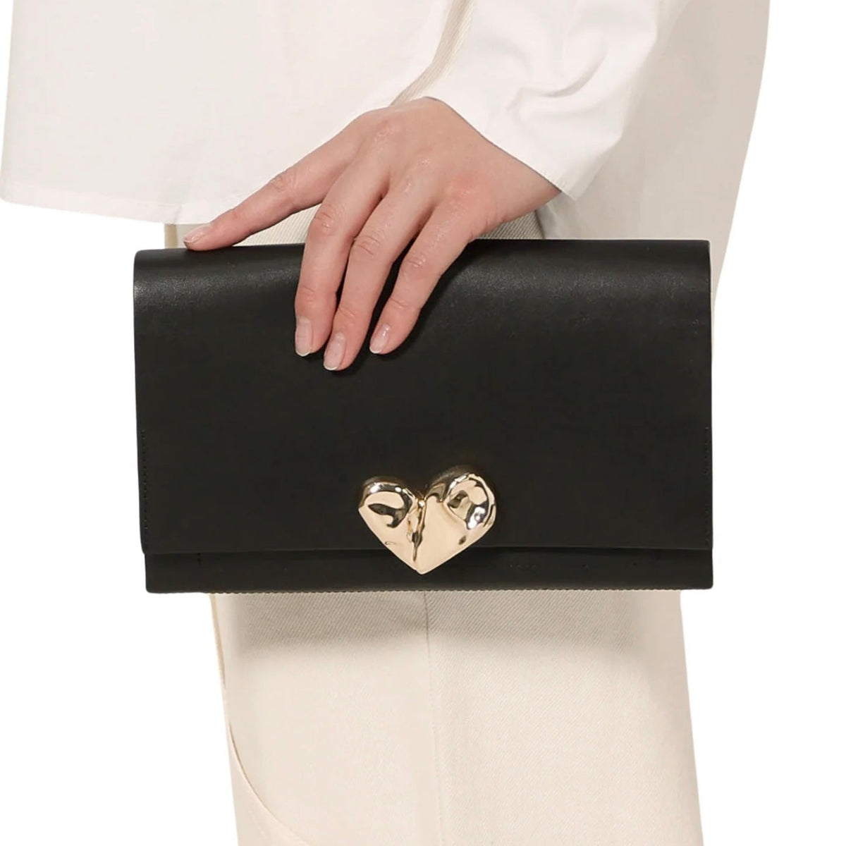 Black clutch with a gold heart clasp held by a person wearing a white outfit.