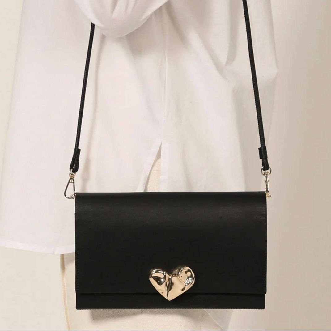 Black handbag with a gold heart detail held by a person wearing a white coat.