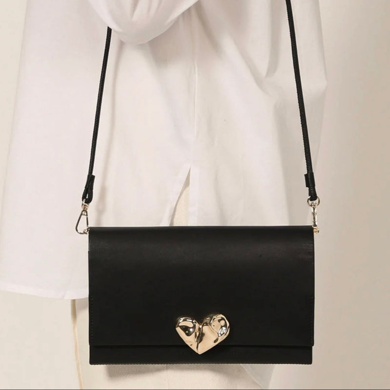 Black handbag with a gold heart detail held by a person wearing a white coat.
