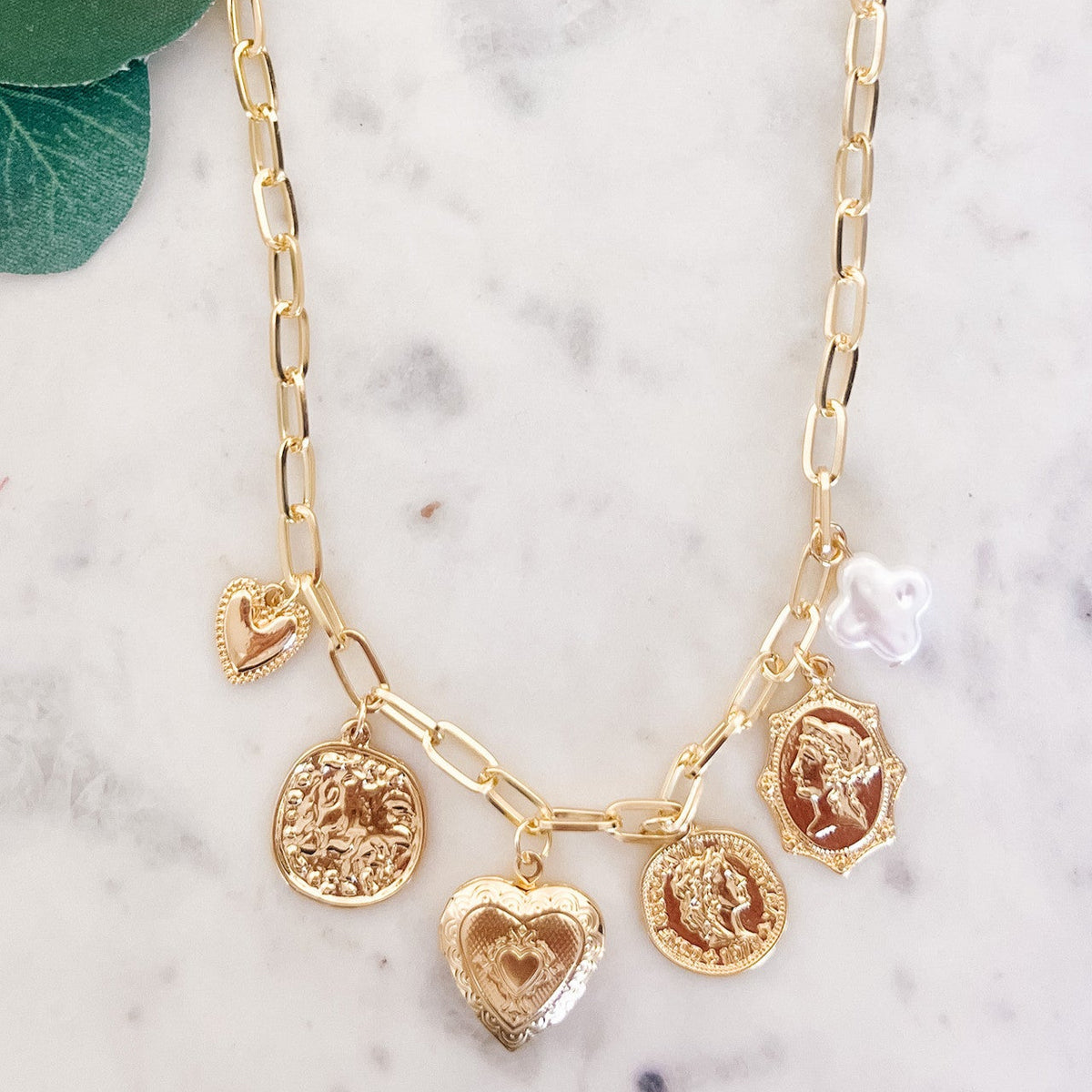 Gold necklace with heart and coin charms on a marble surface with green leaves.