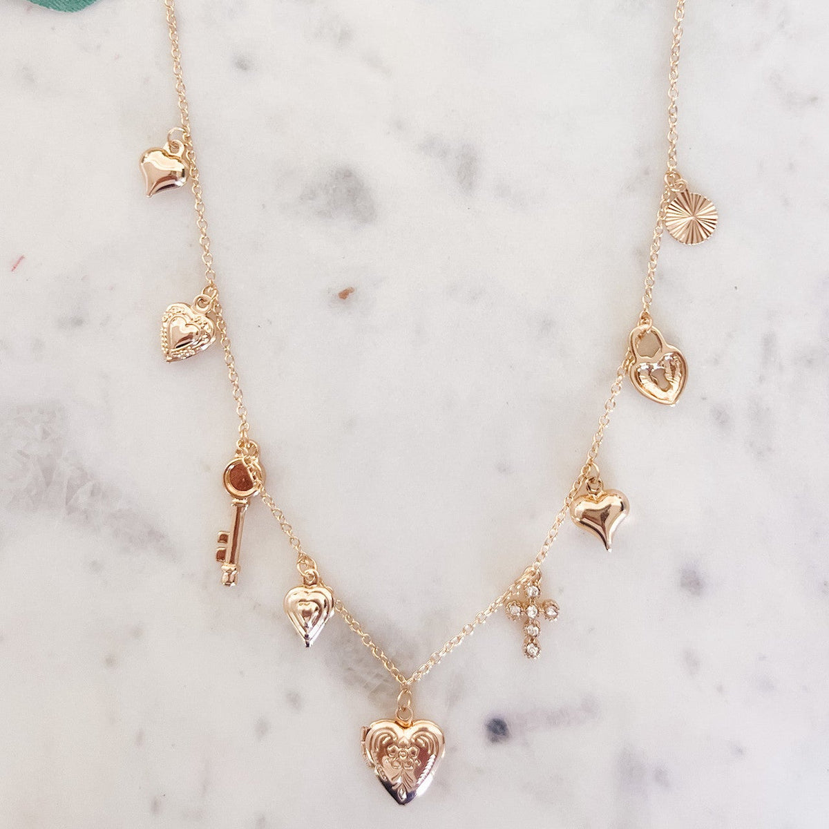 Gold necklace with heart and key pendants on a marble surface with green leaves.