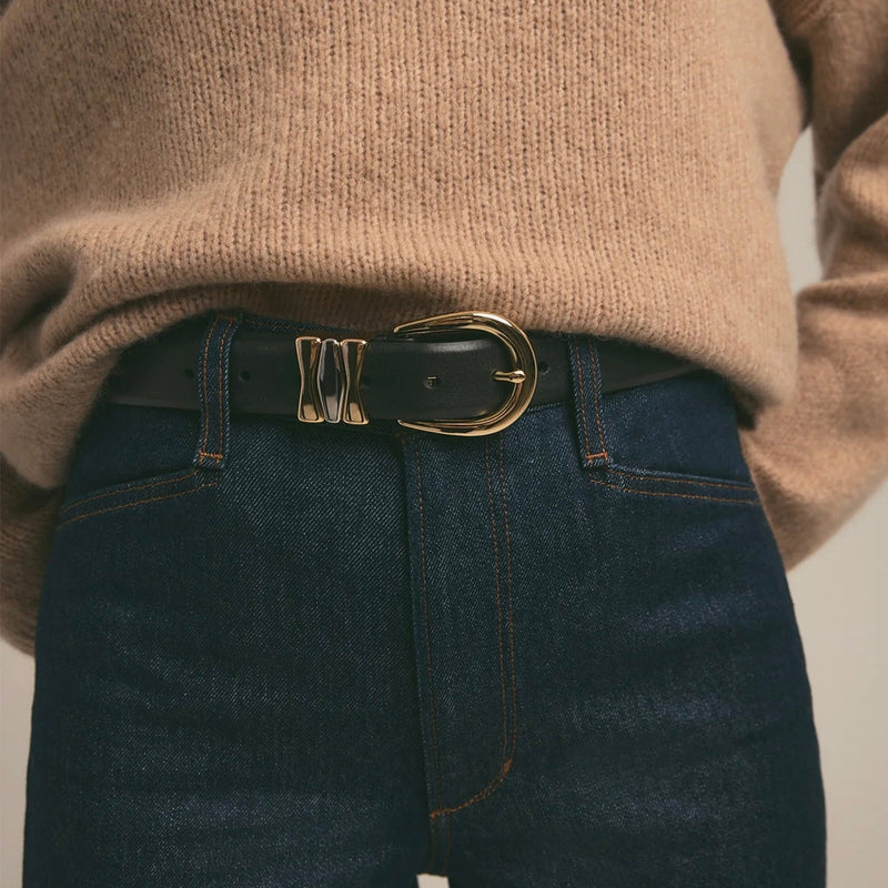 Person wearing a beige sweater and dark jeans with a gold belt buckle on a neutral background