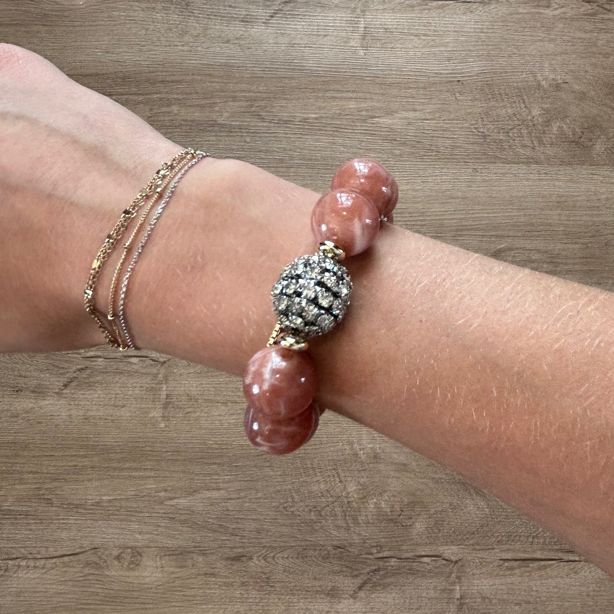 A close-up of a person's wrist wearing a beaded bracelet with pinkish-red stone beads, a textured silver-and-gold central bead, and two delicate gold chain bracelets underneath, set against a wood floor background.