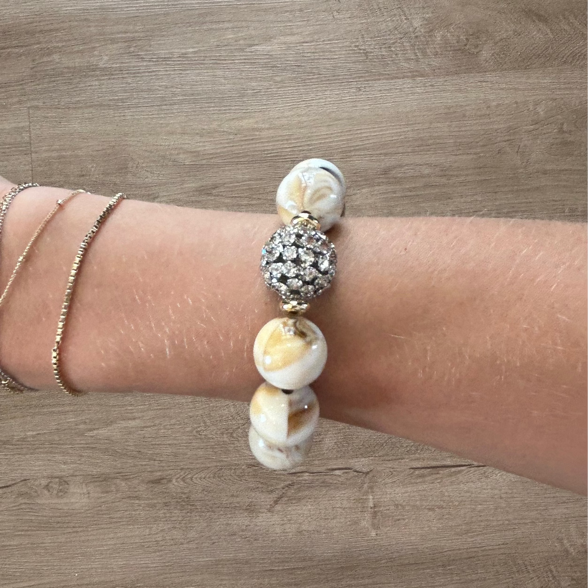 A close-up of a person's wrist wearing a beaded bracelet with white and cream marbled beads, a central silver rhinestone ball, and two delicate gold chain bracelets, set against a light brown wood plank background.