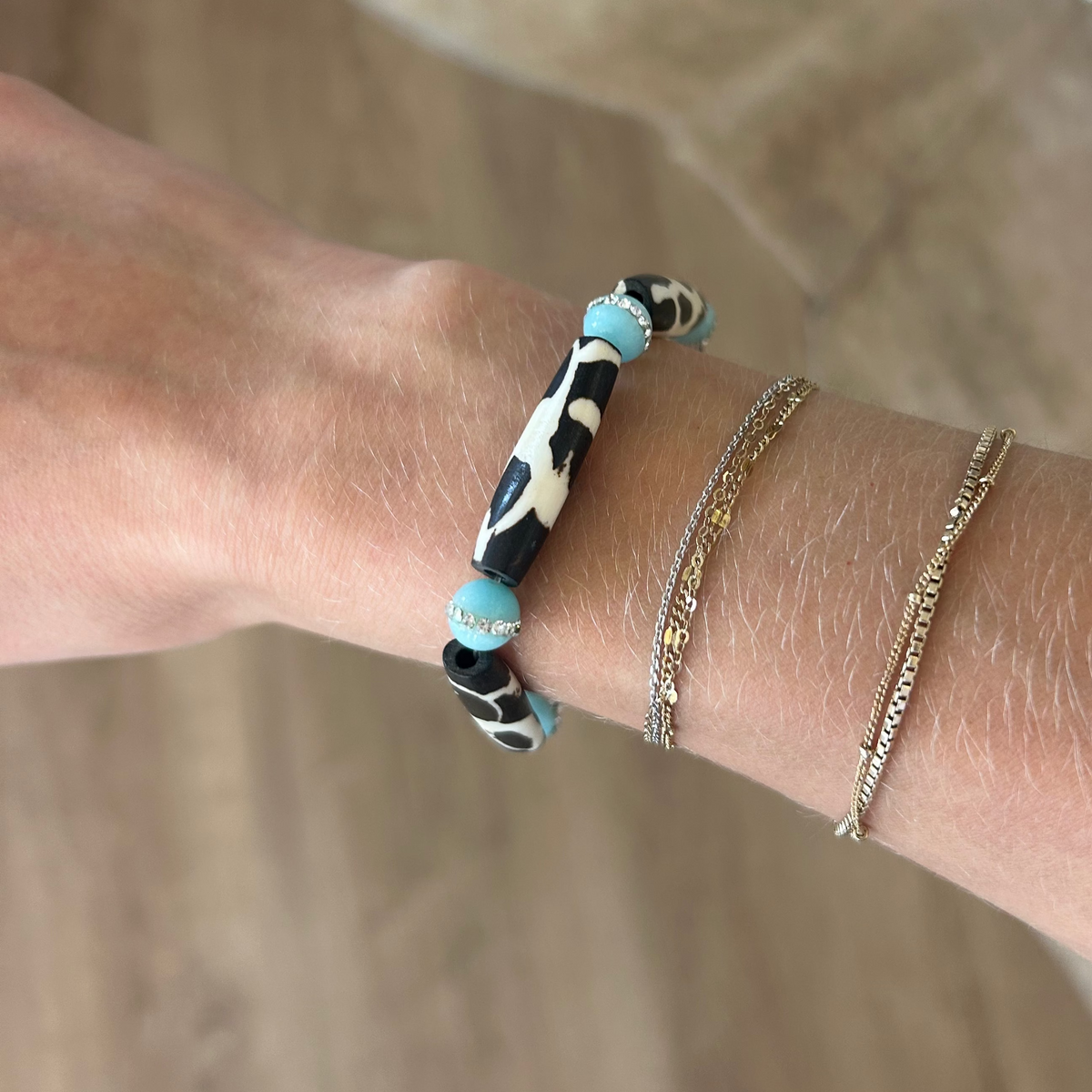 A close-up of a person's wrist wearing three bracelets: a black and white patterned beaded bracelet with turquoise accents, a thin gold chain bracelet, and a textured gold chain bracelet, against a warm brown background.