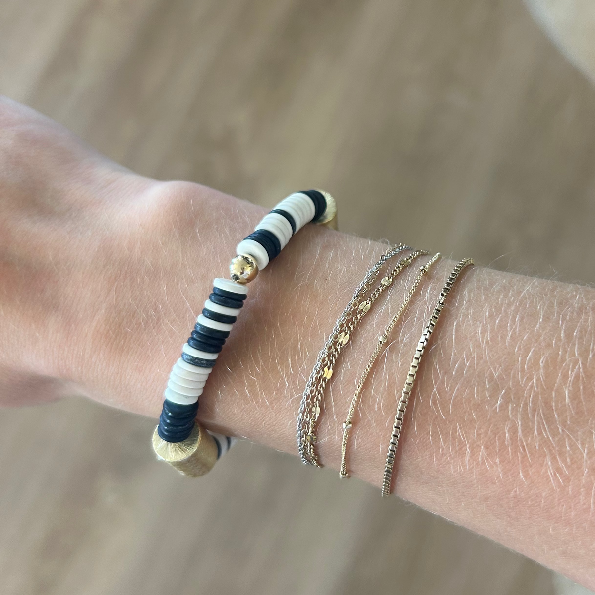 A close-up of a person's wrist wearing multiple bracelets, including a beaded bracelet with navy, white, and gold beads, and two thin gold chain bracelets, against a blurred wood and beige background.