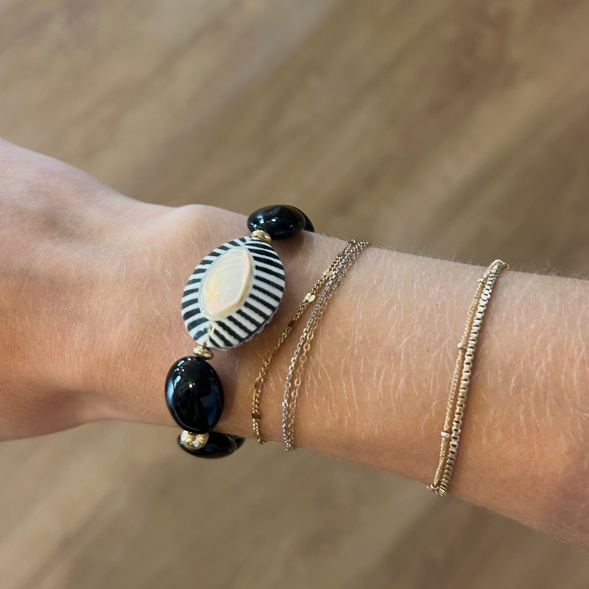 A close-up of a person's wrist wearing three delicate bracelets: a black and white striped oval charm bracelet with black beads, a thin gold chain bracelet, and a textured gold bangle, set against a blurred wooden floor background.
