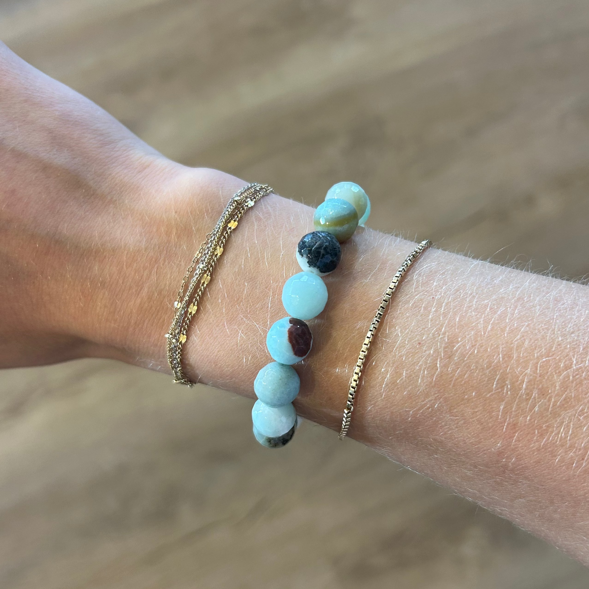 A close-up of a person's wrist wearing multiple beaded and chain bracelets against a blurred wood floor background.