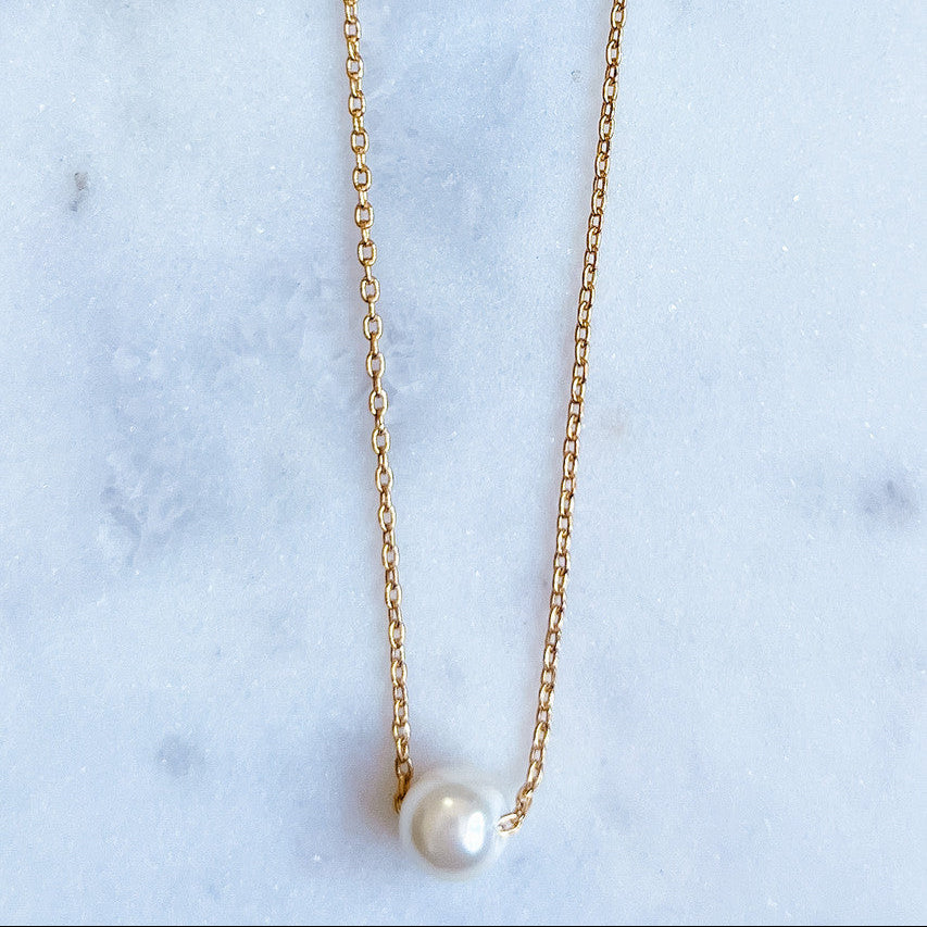 Gold necklace with a pearl pendant on a marble surface with greenery.