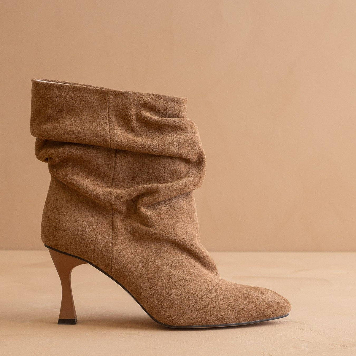 A single tan suede ankle boot with a slouchy draped design and a slim kitten heel, shown in profile against a warm beige background.