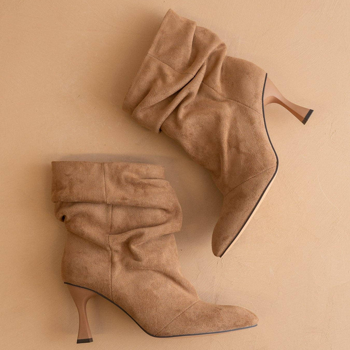 A pair of beige suede ankle boots with mid-height stiletto heels, displayed on a matching beige background.