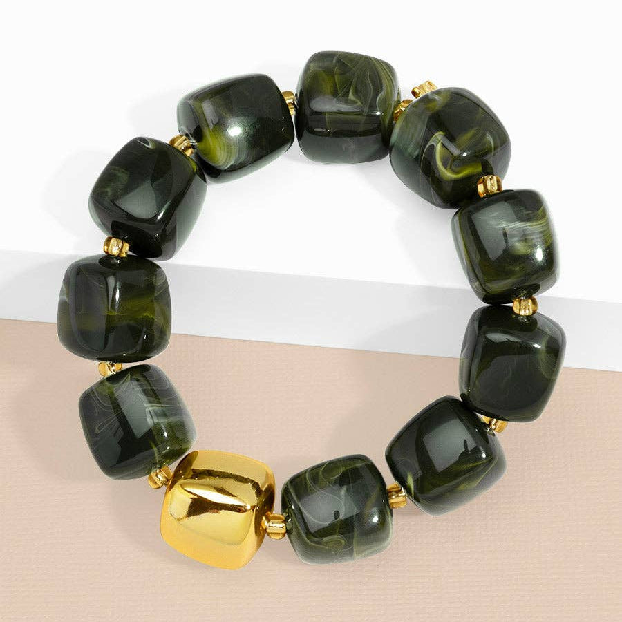 A beaded stretch bracelet featuring glossy dark green and black marbled beads with a prominent shiny gold cube accent bead, displayed on a two-tone white and beige surface.