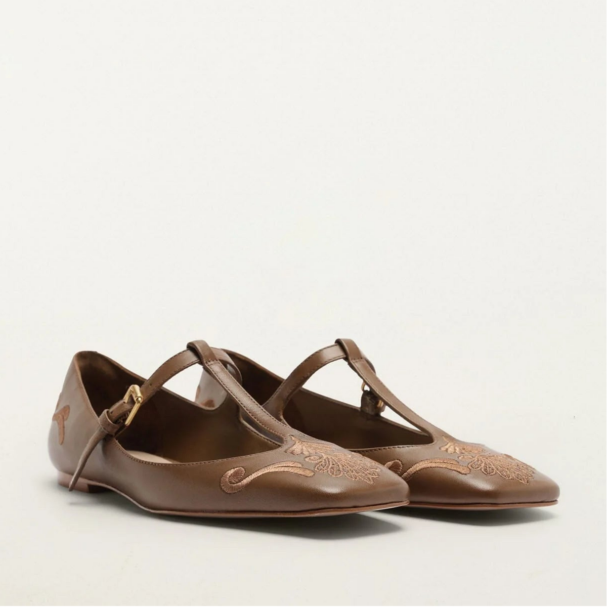 A pair of brown leather ballet flats with T-strap details and decorative floral embroidery on the toes, photographed against a plain off-white background.
