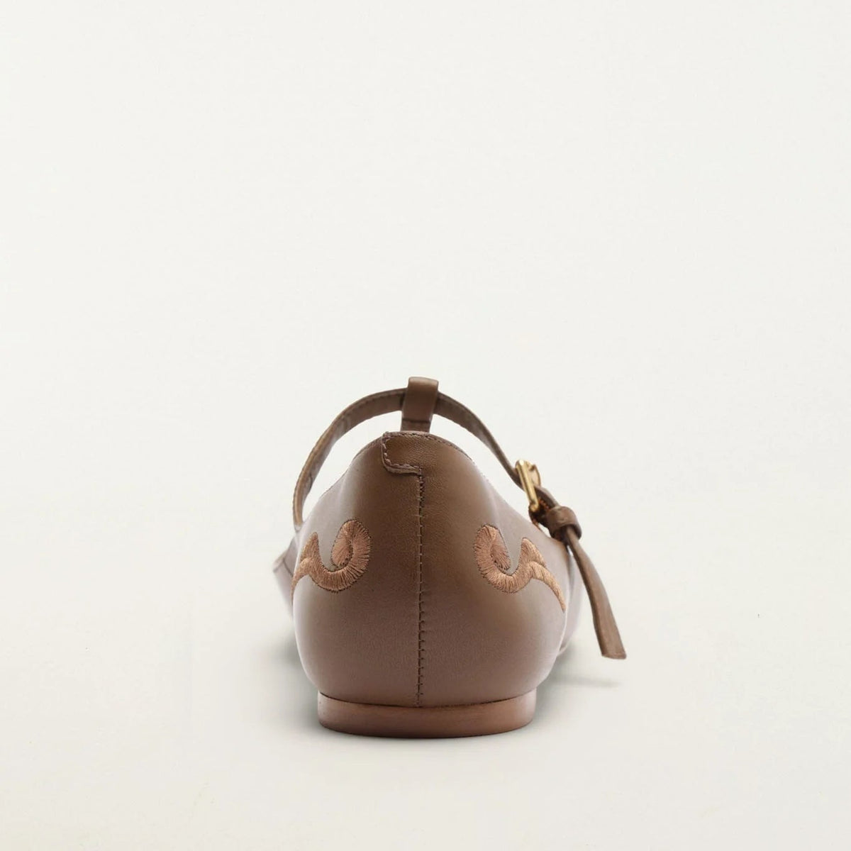 A single brown leather ballet flat shown from the back, featuring decorative stitching, a strap with a tassel, and a small gold-tone buckle, set against a plain white background.