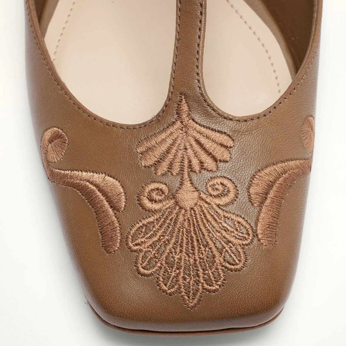 A close-up top-down view of a brown leather flat shoe with ornate matching brown embroidery on the toe area, set against a plain white background.